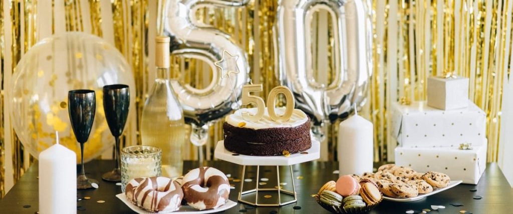 Gâteau de 50ème anniversaire posé sur un socle sur une table avec des gâteaux et du champagne. En fond il y a des ballons en forme de 50.